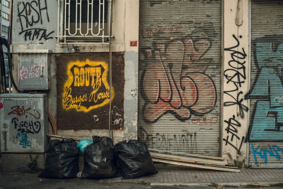 A row of five tightly packed black rubbish bags placed on a concrete sidewalk in front of a weathered building featuring extensive graffiti. The bags appear full and are situated directly beneath a metal utility box with multicolored tags, wires extending from it. To the right, a partially closed metal roller shutter with large, overlapping graffiti tags in red and black is visible, alongside vertical black graffiti on the adjacent wall. On the left, the building's facade exhibits peeling paint and a small, barred window above the bags, with a bright yellow and black painted sign reading 'BOUFFE Burger House' affixed to the wall. The surrounding environment is urban and gritty, emphasizing an exterior scene where private rubbish collection or on-site waste handling by services like those offered by wastecollectionshepherdsbush.co.uk might be necessary to manage clutter in this area.