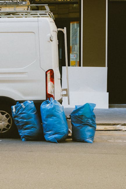 A row of five tightly packed black rubbish bags placed on a concrete sidewalk in front of a weathered building featuring extensive graffiti. The bags appear full and are situated directly beneath a metal utility box with multicolored tags, wires extending from it. To the right, a partially closed metal roller shutter with large, overlapping graffiti tags in red and black is visible, alongside vertical black graffiti on the adjacent wall. On the left, the building's facade exhibits peeling paint and a small, barred window above the bags, with a bright yellow and black painted sign reading 'BOUFFE Burger House' affixed to the wall. The surrounding environment is urban and gritty, emphasizing an exterior scene where private rubbish collection or on-site waste handling by services like those offered by wastecollectionshepherdsbush.co.uk might be necessary to manage clutter in this area.