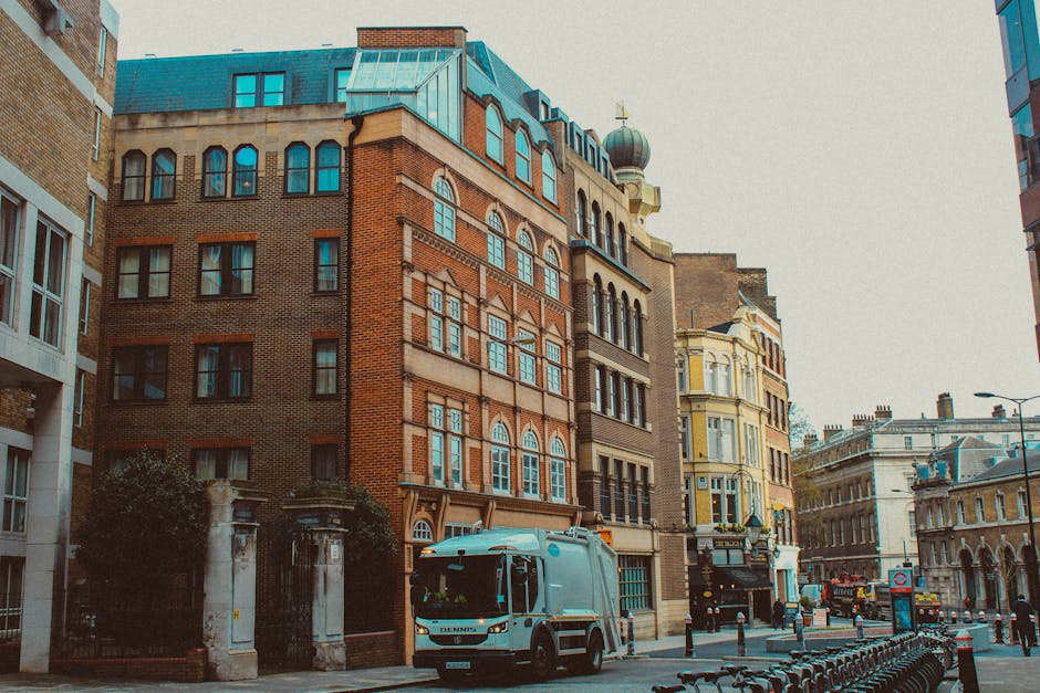 A street scene featuring a collection of older, multi-story buildings with varied architectural styles, including brick facades, arched windows, and decorative elements, located in an urban area with narrow pavement. In the foreground, a light blue rubbish collection vehicle is parked on the street, positioned near a waist-high metal barrier that separates the cycling lane from the traffic lane. The vehicle appears to be a compact, modern waste disposal truck with a smooth, metallic finish and dark tinted windows, indicating ongoing rubbish or waste collection services. Surrounding buildings include a terraced structure with a rounded turret and a green-domed tower, alongside others with light-colored stucco and ornate window frames, suggesting a mixture of historic and contemporary architecture. The street backdrop features additional vehicles, street lamps, and signage. Natural daylight illuminates the scene, with a slight overcast sky creating diffuse lighting, aligning with the context of private waste collection or on-site rubbish clearance services in a busy city environment, with no visible waste or debris in sight.
