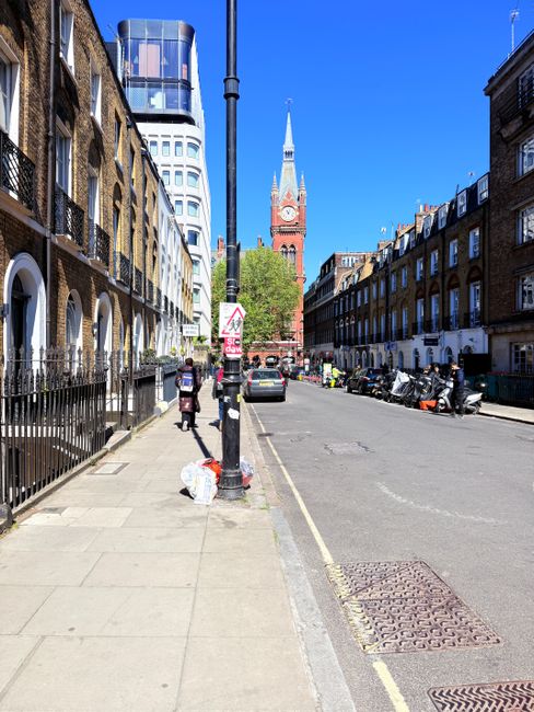 A wide urban street scene in daylight showing a row of brick terraced buildings on the left with iron railings and a modern glass-fronted office building at the far end, and a line of parked motorbikes on the right. In the foreground, a black streetlamp pole stands on the pavement, with a plastic bag of rubbish placed nearby on the sidewalk. The street is lined with several cars parked parallel to the curb, and pedestrians are visible walking along the pavement. Central in the background is a tall, red-brick clock tower with a spire, part of a historic building, set against a bright blue sky. The scene appears to be in a city area where private waste collection or rubbish removal services like those offered by wastecollectionshepherdsbush.co.uk could operate for efficient on-site or alternative waste handling, captured in a clear, detailed manner suitable for accessibility and SEO purposes.
