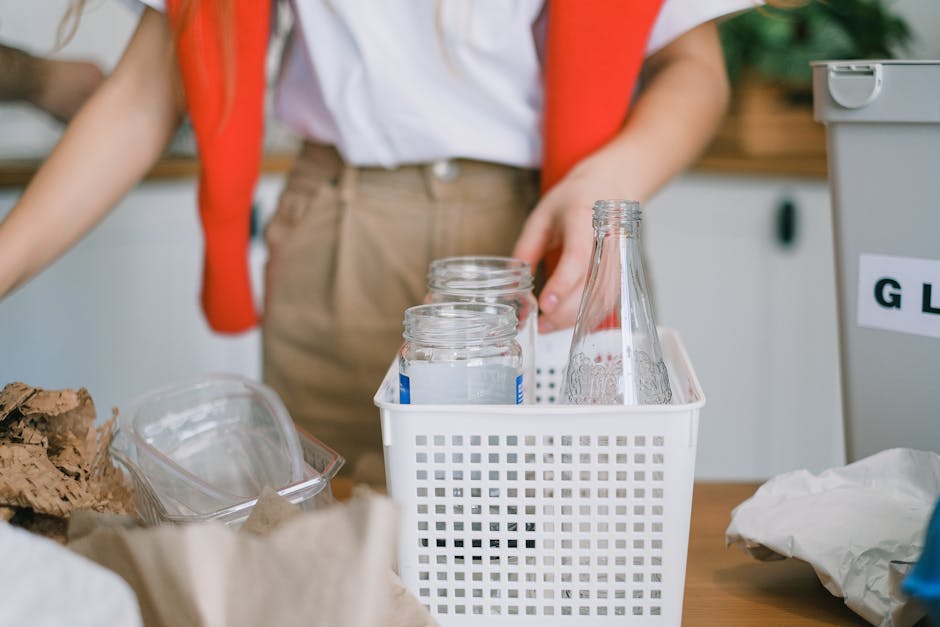 In the foreground, a close-up view shows a white plastic basket with a grid-like pattern, holding several empty glass bottles and jars, some with metal screw caps and others without. The bottles and jars are transparent, with smooth surfaces and varying shapes, positioned upright within the basket. To the left, there is a partially visible clear plastic container filled with crumpled brown paper or cardboard packaging material. The background features a person wearing beige trousers and a white shirt, with a red item draped over their shoulders, standing behind a cluttered workspace or table. Visible on the right side is a grey recycling bin or container with a label, likely indicating glass or general waste, placed in an indoor environment that suggests an organized space for waste sorting or collection. The scene is well-lit, emphasizing the different textures and materials of the bottles, packaging, and containers, and subtly illustrates a setting related to designated waste handling, such as glass recycling or private disposal.
