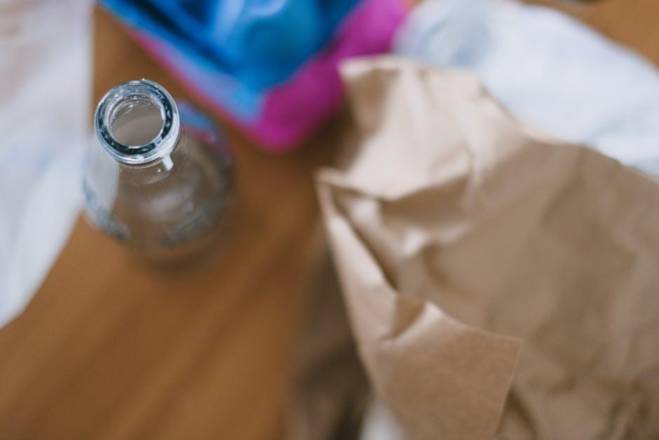 A clear glass bottle with an open neck and no label is positioned on a wooden surface, with the opening facing upward. Behind it, there is a partially visible cardboard box or bag and a colorful item that appears to be a container or packaging, both slightly out of focus. The environment suggests an indoor setting, possibly a kitchen or storage area, with natural or ambient lighting. The scene relates to waste or rubbish collection, as the glass bottle and packaging materials are typically disposed of during cleaning or decluttering processes, which companies like wastecollectionshepherdsbush.co.uk often handle as part of private rubbish removal or on-site clearance services. The materials show glass with a smooth, reflective surface and the cardboard with a crinkled texture, adhering to the typical appearance of waste items prepared for recycling or disposal.